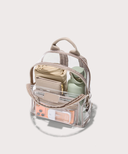 Clear bag with beige straps and a front pocket holding everyday items on a white background. Stadium compliant.