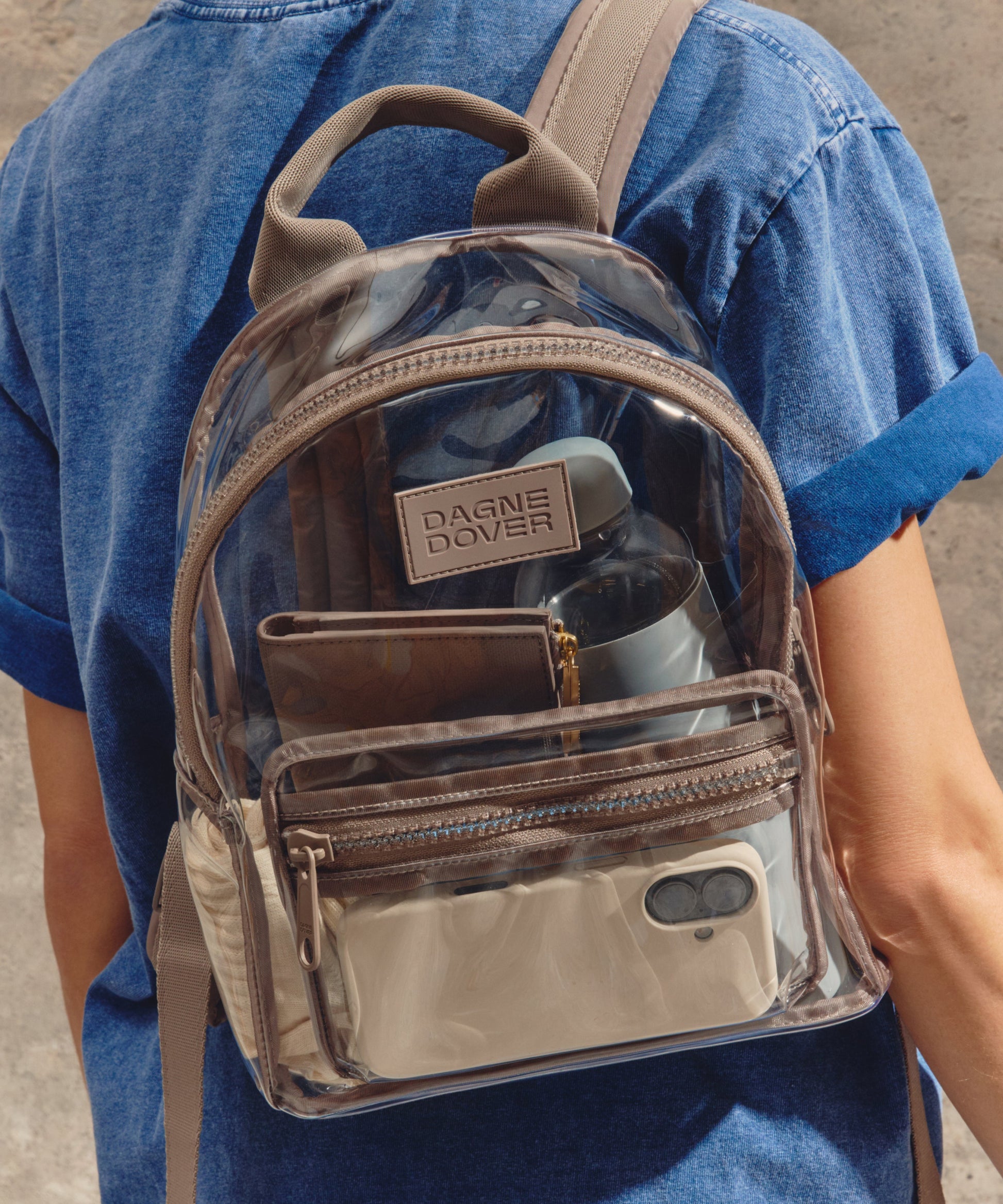 Person wearing a transparent backpack with beige straps and a visible brand logo.Stadium compliant.