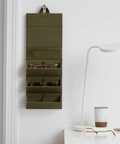Dagne Dover Remi foldable glasses holder in dark green hung up and holding glasses by a desk.