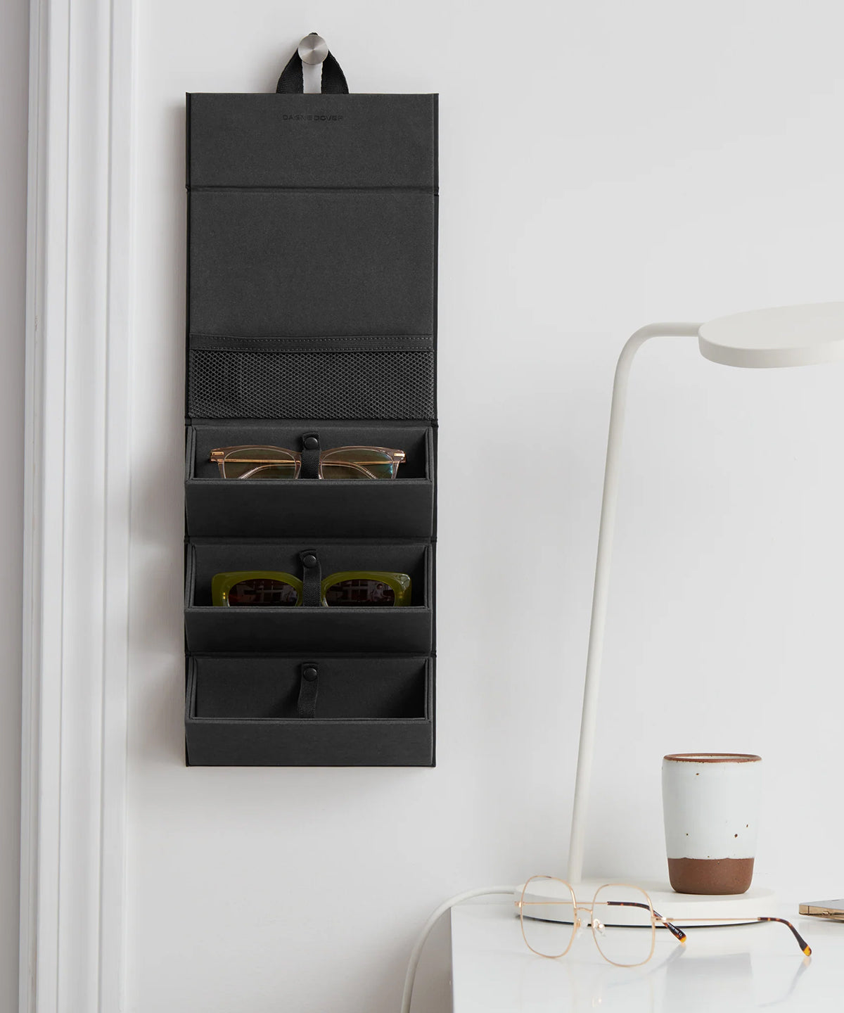 Dagne Dover Remi foldable glasses holder in spice red hung up and holding glasses by a desk.